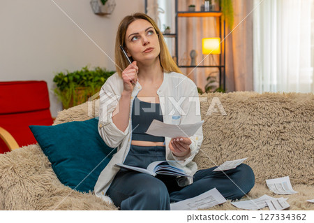 Adult woman reviewing bills and writing expenses in notebook at home, focused concentrated on budget Adult woman reviewing bills and writing expenses in notebook at home, focused concentrated on budget 127334362
