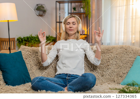 Adult woman meditating with closed eyes at home, listening to music through headphones on sofa 127334365