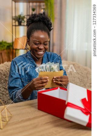 African American young woman opens birthday gift box with envelope of cash dollars sitting at home African American young woman opens birthday gift box with envelope of cash dollars sitting at home 127334369