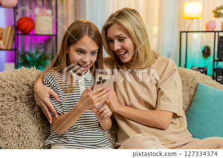 Child girl kid playing smartphone game with mother on home sofa celebrating win cheering with joy Child girl kid playing smartphone game with mother on home sofa celebrating win cheering with joy 127334374