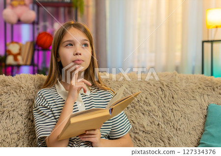 Child girl kid reading interesting book with smile enjoying the story while sitting alone on sofa Child girl kid reading interesting book with smile enjoying the story while sitting alone on sofa 127334376