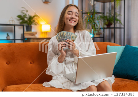 Rich happy young woman counting money cash using laptop calculate domestic bills sitting on sofa Rich happy young woman counting money cash using laptop calculate domestic bills sitting on sofa 127334395