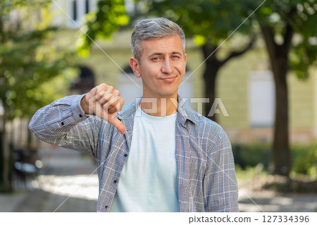 Upset mature man showing thumb down sign gesture, expressing dissatisfied bad work on city street 127334396
