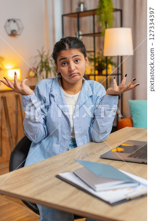 Uncertain young Indian woman freelancer shrugging shoulders don't know how to work at office table 127334397