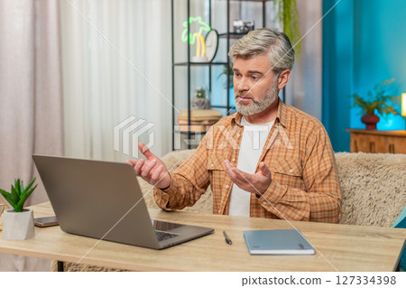 Mature man sitting at table with laptop making serious business video call conversation at home 127334398