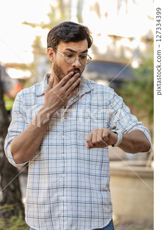 Shocked man worrying about being punctual with anxiety checking time on watch walking on city street 127334399