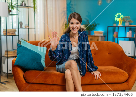 Woman smiling friendly at camera and waving hands gesturing hello, hi, greeting talking at home 127334406