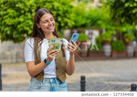 Happy Caucasian woman counting money euro cash, using smartphone calculator app on city street Happy Caucasian woman counting money euro cash, using smartphone calculator app on city street 127334408