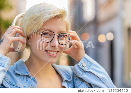 Portrait of smiling young woman in glasses taking off wireless headphones while looking at camera 127334420