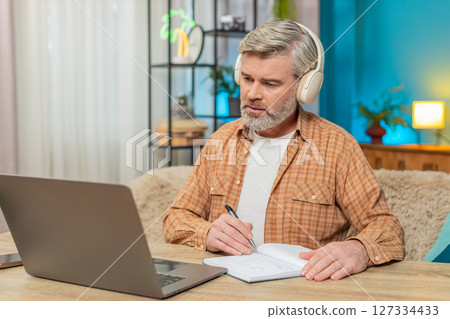 Mature man with laptop and headphones, listening to podcast making notes education study at home 127334433