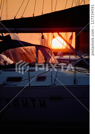 A yacht club at sunset, the big sun in the background, the sail is mounted on the mainsail, sailboat is moored to the berths, thick ropes, reflections in the glossy sides of boats 127334591