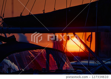 A yacht club at sunset, the big sun in the background, the sail is mounted on the mainsail, sailboat is moored to the berths, thick ropes, reflections in the glossy sides of boats A yacht club at sunset, the big sun in the background, the sail is mounted on the mainsail, sailboat is moored to the berths, thick ropes, reflections in the glossy sides of boats 127334610
