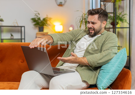 Upset young Caucasian young man sitting on sofa at home using laptop having anxiety and stress Upset young Caucasian young man sitting on sofa at home using laptop having anxiety and stress 127334616