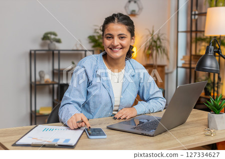 Indian woman working with financial graphs, calculate taxes for charts and reports at desk at home 127334627