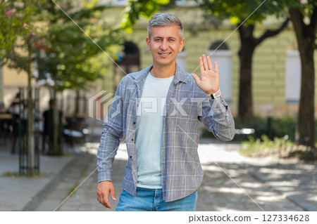 Cheerful mature man tourist smiling at camera, waving hands gesturing hello standing on street Cheerful mature man tourist smiling at camera, waving hands gesturing hello standing on street 127334628