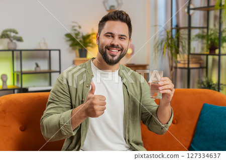 Smiling Caucasian man sitting on sofa leading healthy lifestyle, drinking a glass of pure water 127334637
