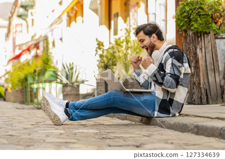 Happy man using laptop celebrating win good message news, lottery jackpot victory sitting on street Happy man using laptop celebrating win good message news, lottery jackpot victory sitting on street 127334639