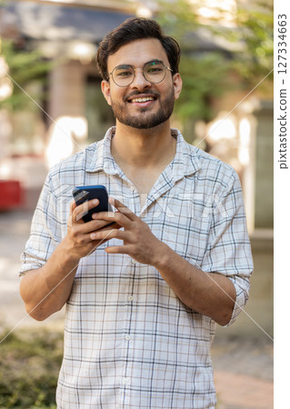 Hispanic young man using smartphone text messages browsing internet smiling at camera outdoors 127334663