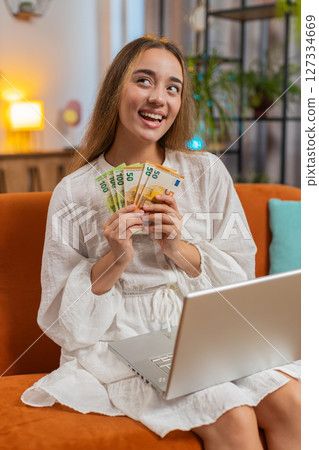 Rich happy young woman counting money euro cash using smartphone calculator sitting at home 127334669