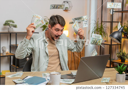 Successful young man freelancer using laptop celebrate success falling money at home office table Successful young man freelancer using laptop celebrate success falling money at home office table 127334745