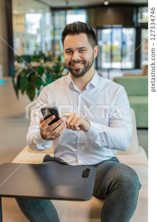 Smiling Caucasian businessman using smartphone typing text messages browsing news in office lobby 127334746