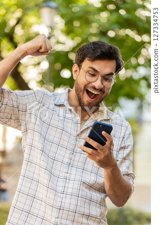 Hispanic young man using smartphone celebrating win good message news clenching fist on city street Hispanic young man using smartphone celebrating win good message news clenching fist on city street 127334753