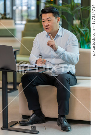 Chinese businessman writing down notes while attending online video class in modern office lobby Chinese businessman writing down notes while attending online video class in modern office lobby 127334754