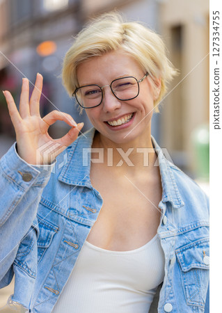 Portrait of attractive young millennial woman looking at camera showing hand OK sign on city street 127334755
