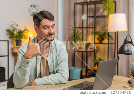 Happy Caucasian man looking approvingly at camera showing thumbs up sitting at home office table 127334758