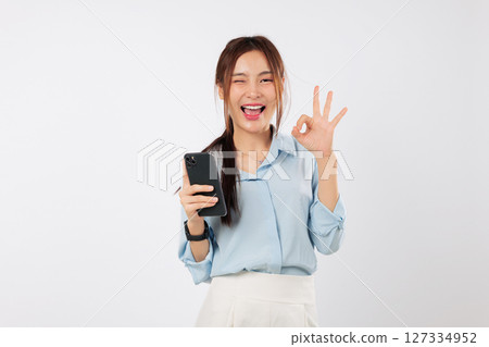 Excited Young Asian woman smiles, holding a mobile phone and giving an ok gesture sign. She is happy showing her smart phone. social media network isolated studio white background Excited Young Asian woman smiles, holding a mobile phone and giving an ok gesture sign. She is happy showing her smart phone. social media network isolated studio white background 127334952