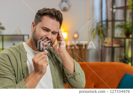 Man suffering from headache taking pill treatment sitting on sofa in living room at home Man suffering from headache taking pill treatment sitting on sofa in living room at home 127334976