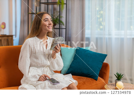 Rich happy young woman counting money cash using smartphone calculator sitting on sofa at home Rich happy young woman counting money cash using smartphone calculator sitting on sofa at home 127334978