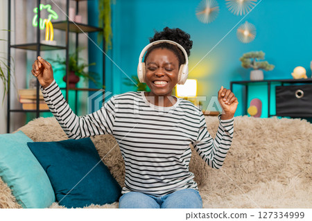 African American woman girl enjoying listening music, dancing on sofa, wireless headphones at home African American woman girl enjoying listening music, dancing on sofa, wireless headphones at home 127334999