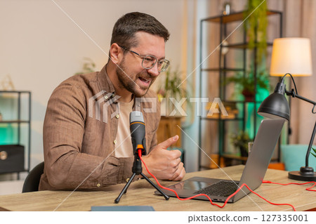 Happy young Caucasian man influencer speaking into microphone and laptop webcam at home office table 127335001