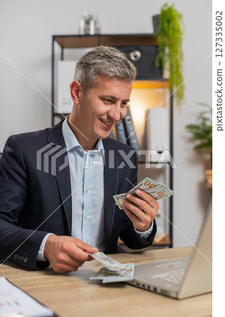 Caucasian young businessman in suit working on laptop PC counting money cash at home office desk 127335002