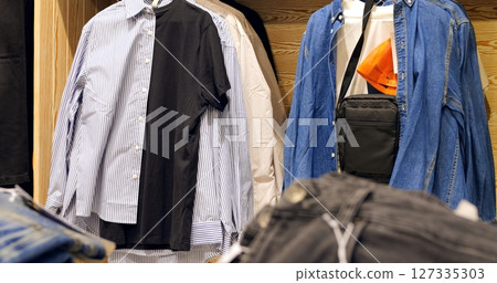 Fashion retail display presenting variety of clothing items, shirts, jeans, and black shoulder bag hanging neatly on store racks, showcasing contemporary casual menswear selection 127335303