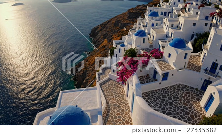 Sunlight reflects on the aegean sea near the whitewashed houses and blue domed churches of oia village, santorini, greece, with blooming bougainvillea draping over the walls 127335382