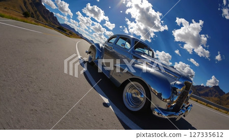 Polished chrome vintage car parked on a scenic mountain road gleams under the sunlight, reflecting the surrounding landscape and cloudy sky 127335612