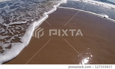 Sparkling white foamy waves gently washing over golden sandy coastline, creating mesmerizing natural patterns under bright sunlight along serene oceanfront landscape 127335657