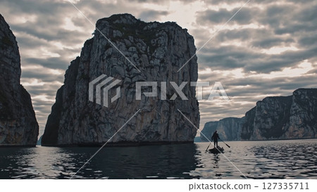 Rugged rocky cliffs rising from dark waters under overcast sky, with lone paddleboarder navigating calm seascape during serene twilight journey 127335711