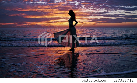 Golden hour casting dramatic light on elegant model standing on wet sand, reflecting silhouette against ocean waves under vibrant sunset sky 127335712