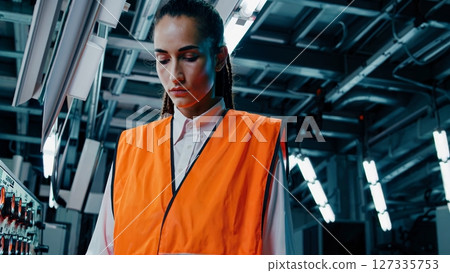 Professional female engineer wearing an orange safety vest, meticulously inspecting machinery and equipment in a modern industrial factory, ensuring smooth operations and safety protocols 127335753