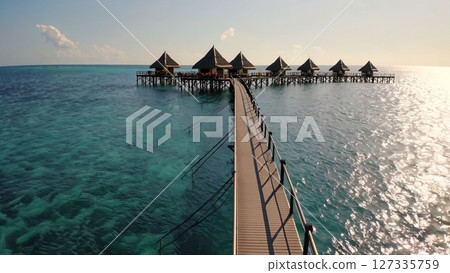 Wooden walkway connects overwater bungalows with thatched roofs, offering a luxurious and tranquil retreat in crystal clear turquoise waters, creating an idyllic tropical paradise 127335759