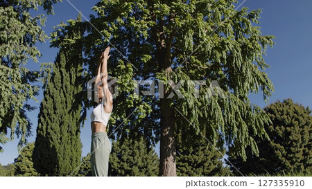 Black female yogi meditating outdoors, performing stretching pose with raised arms, embracing peaceful mindfulness near verdant greenery 127335910