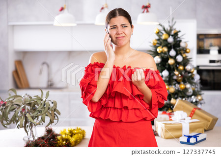 Distressed young European woman in red dress speaking on smartphone next to wrapped gifts and Christmas tree before holidays at flat 127337745
