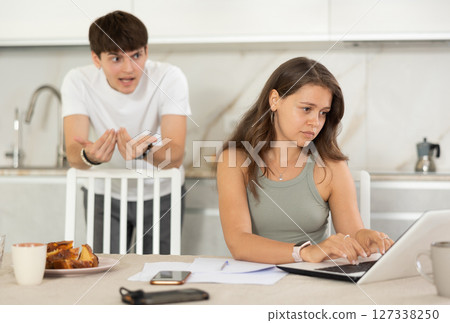 Young girl sitting at the table while her boyfriend is quarreling with her standing behind 127338250