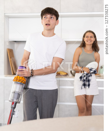 Guy vacuum-cleaning in the kitchen while communicating with his girlfriend drying dishes with towel Guy vacuum-cleaning in the kitchen while communicating with his girlfriend drying dishes with towel 127338275