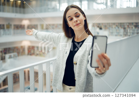 Selfie, library and student with smartphone for social media, video call or influencer promotion of scholarship, knowledge and education. Live streaming, blog update and woman show college bookshelf 127341094