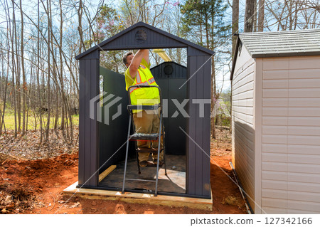 Worker in reflective vest puts finishing touches at storage shelter while standing on ladder Worker in reflective vest puts finishing touches at storage shelter while standing on ladder 127342166
