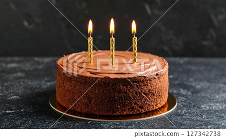 Chocolate Cake with Candles: A delectable chocolate cake, adorned with three lit golden candles, sits majestically on a dark background. 127342738